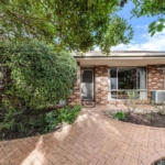 A single-story brick house with a front porch, surrounded by trees and shrubs, awaits its new owners. Featuring a screen door entrance and a large window on the right, this charming home, highlighted by Yes Property Agents, boasts a walkway paved with herringbone-patterned bricks. From Yes property agents website